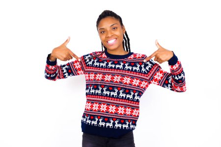 Foto de ¡Escoge! Mujer afroamericana segura, segura de sí misma y carismática que usa suéter de Navidad contra la pared blanca promoviéndose a sí misma como queriendo un papel sonriente ampliamente y apuntando al cuerpo. - Imagen libre de derechos