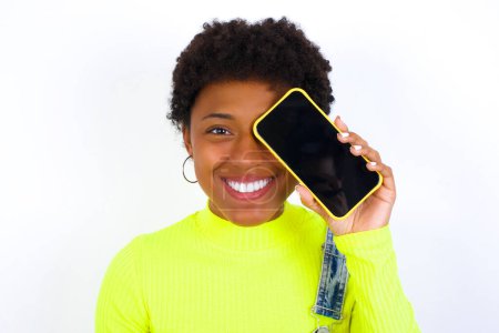 Foto de Joven mujer afroamericana con el pelo corto con denim en general contra la pared blanca sosteniendo teléfono inteligente moderno que cubre un ojo mientras sonríe - Imagen libre de derechos