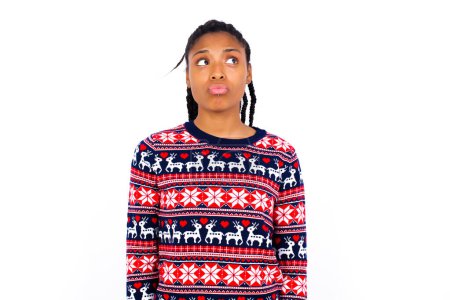 Téléchargez les photos : Femme afro-américaine portant un pull de Noël contre un mur blanc regarde pensivement de côté, planifie des actions après l'université, imagine ce qu'il faut faire Pense à nouveau projet. - en image libre de droit