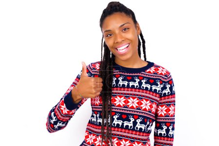 Photo for African American woman wearing Christmas sweater against white wall giving thumb up gesture, good Job! Positive human emotion facial expression body language. - Royalty Free Image