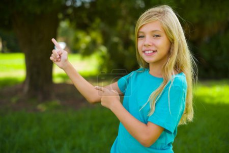Photo for Portrait of beautiful caucasian little kid girl wearing blue t-shirt standing outdoor in the park pointing with finger - Royalty Free Image