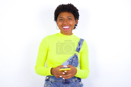 Photo for Pleasant looking happy young African American woman with short hair wearing denim overall against white using telephone and typing message - Royalty Free Image
