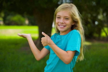 Photo for Portrait of beautiful caucasian little kid girl wearing blue t-shirt standing outdoor in the park advertising something on empty space - Royalty Free Image