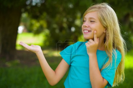 Téléchargez les photos : Portrait de belle petite fille caucasienne vêtue de t-shirt bleu debout en plein air dans le parc annonçant quelque chose sur l'espace vide - en image libre de droit