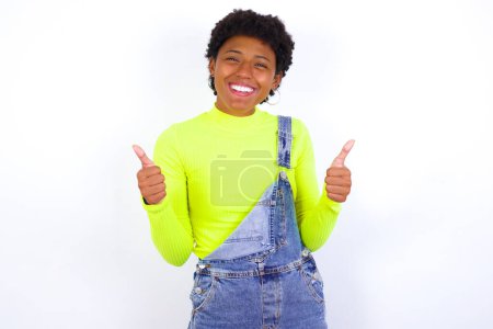 Photo for Optimistic young African American woman with short hair wearing denim overall against white wall showing thumbs up with positive emotions. Quality and recommendation concept. - Royalty Free Image