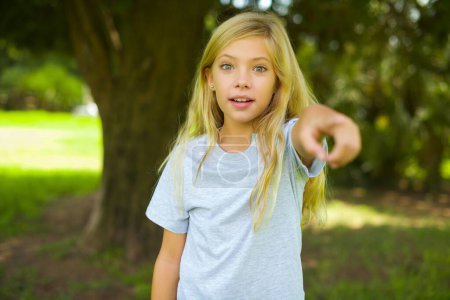 Foto de Emocionado positiva niña caucásica usando camiseta blanca de pie al aire libre en los puntos del parque dedo índice directamente en usted, ve algo muy divertido. Wow, increíble - Imagen libre de derechos