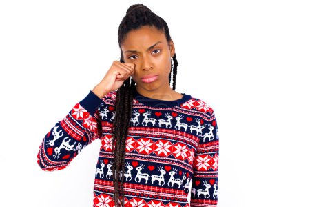 Photo for Unhappy African American woman wearing Christmas sweater against white wall crying while posing at camera wiping tears with hand. - Royalty Free Image