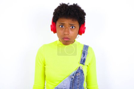 Téléchargez les photos : Serieux mécontent jeune afro-américaine aux cheveux courts portant le denim dans l'ensemble contre le mur blanc semble perplexe à la caméra étant en colère porte casque stéréo écoute de la musique tout en marchant dans la rue - en image libre de droit