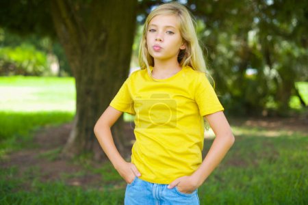 Foto de Retrato de encantadora niña caucásica divertida usando camiseta amarilla de pie al aire libre enviando beso de aire - Imagen libre de derechos