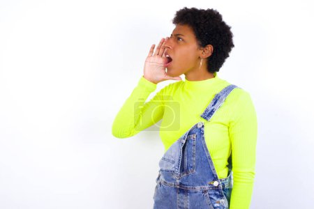 Photo for Young African American woman with short hair wearing denim overall against white wall profile view, looking happy and excited, shouting and calling to copy space. - Royalty Free Image