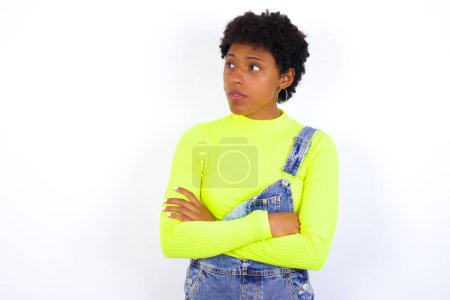 Téléchargez les photos : Serious jeune femme afro-américaine avec les cheveux courts portant le denim dans l'ensemble contre le mur blanc garde les mains croisées se tient dans la pose réfléchie concentrée quelque part - en image libre de droit