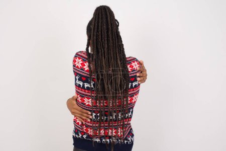Téléchargez les photos : Femme afro-américaine portant un pull de Noël contre un mur blanc s'embrassant heureux et positif à partir de l'arrière. Amour de soi et soin de soi. - en image libre de droit