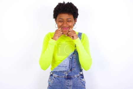 Photo for Pleased young African American woman with short hair wearing denim overall against white wall with closed eyes keeps hands near cheeks and smiles tenderly imagines something very pleasant - Royalty Free Image