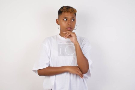 Photo for Shot of contemplative thoughtful Young african-american woman wearing white t-shirt keeps hand under chin, looks thoughtfully upwards, dressed in casual clothes, poses over white background with free space for your text - Royalty Free Image