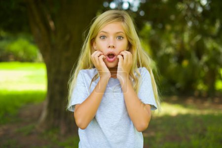 Sprachloses kaukasisches kleines Mädchen in weißem T-Shirt, das draußen im Park steht und die Hände in der Nähe des geöffneten Mundes hält, reagiert auf schockierende Nachrichten und starrt verwundert in die Kamera