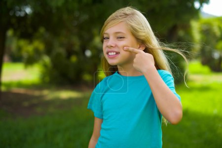 Foto de Niña caucásica usando camiseta azul de pie al aire libre con la cara feliz sonriendo y sosteniendo el dedo en la barbilla - Imagen libre de derechos