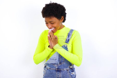 Photo for Overjoyed young African American woman with short hair wearing denim overall against white wall laughs joyfully and keeps palms pressed together hears something funny - Royalty Free Image