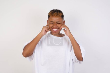 Téléchargez les photos : Arrête de faire ce bruit ennuyeux ! Headshot of unhappy stressé jeune femme afro-américaine portant un t-shirt blanc faisant face à l'inquiétude, bouchant les oreilles avec les doigts, irrité par le bruit fort provenant de voisins qui vivent au-dessus d'elle. - en image libre de droit