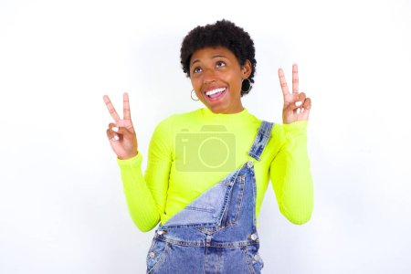 Photo for Isolated shot of cheerful young African American woman with short hair wearing denim overall against white wall makes peace or victory sign with both hands, feels cool. - Royalty Free Image
