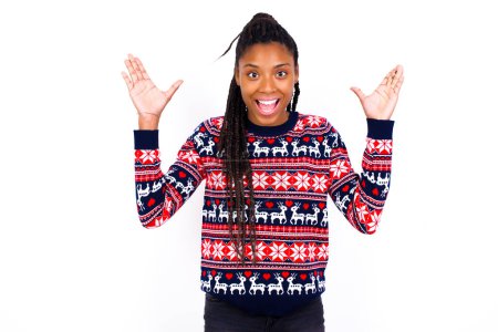Téléchargez les photos : Femme afro-américaine portant un pull de Noël contre un mur blanc levant les mains, ayant les yeux pleins de bonheur se réjouissant de ses grandes réalisations. Réalisation, concept de réussite. - en image libre de droit