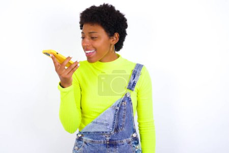 Foto de Mujer joven afroamericana sonriente con el pelo corto usando denim en general contra la pared blanca enviando un mensaje de voz en su teléfono inteligente. Comunicación y concepto de nuevas tecnologías. - Imagen libre de derechos