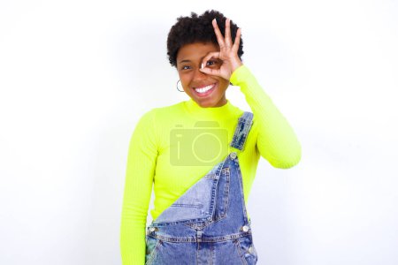 Foto de Joven mujer afroamericana con el pelo corto con denim en general contra la pared blanca con la cara feliz sonriendo haciendo signo bien con la mano en el ojo mirando a través del dedo. - Imagen libre de derechos