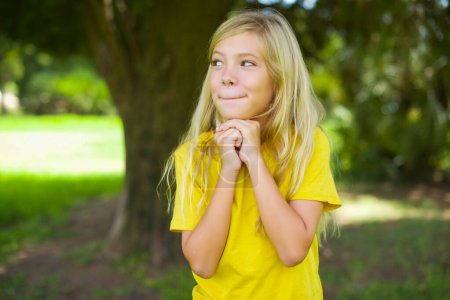 Photo for Curious caucasian little girl wearing yellow t-shirt standing outdoors keeps hands under chin bites lips and looks with interest aside. - Royalty Free Image
