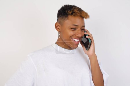 Foto de Lifestyle Concept - Retrato de una hermosa joven afroamericana con una camiseta blanca hablando por teléfono móvil con un amigo. Fondo de estudio blanco. Copiar espacio. - Imagen libre de derechos