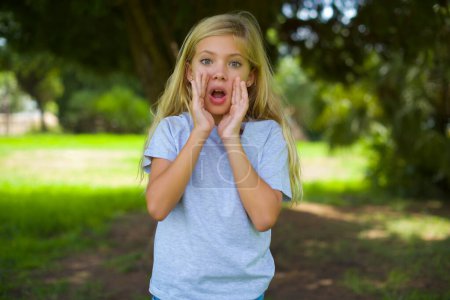 caucasian little kid girl wearing white t-shirt standing outdoor in the park touching forehead, hears something surprising, glad receive good news, feels relieved. Almost got in trouble.