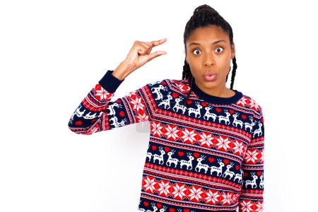 Téléchargez les photos : Choquée femme afro-américaine portant un pull de Noël contre un mur blanc montre quelque chose de peu avec les mains, démontre la taille, ouvre la bouche de surprise. Concept de mesure. - en image libre de droit