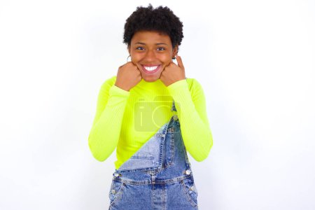 Foto de Feliz joven mujer afroamericana con el pelo corto con denim en general contra la pared blanca mantiene los puños en las mejillas sonríe ampliamente y tiene expresión positiva de estar de buen humor - Imagen libre de derechos