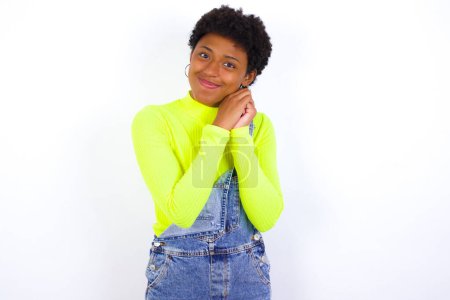 Photo for Charming serious young African American woman with short hair wearing denim overall against white wall keeps hands near face smiles tenderly at camera - Royalty Free Image