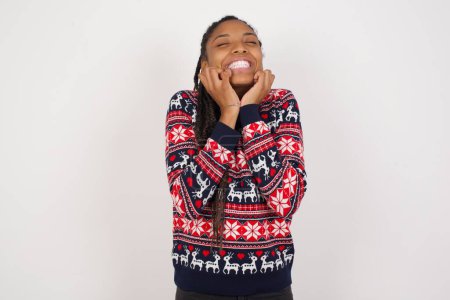 Photo for Portrait of African American woman wearing Christmas sweater against white wall being overwhelmed, expressing excitement and happiness with closed eyes and hands near face. - Royalty Free Image