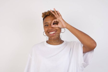 Téléchargez les photos : Belle jeune femme afro-américaine portant un t-shirt blanc avec un visage heureux souriant faisant signe ok avec la main sur les yeux regardant à travers le doigt. Porter des vêtements décontractés et debout sur un mur blanc. - en image libre de droit