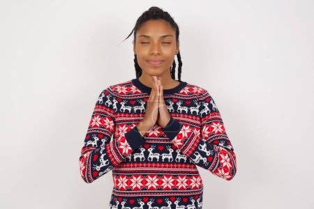 Téléchargez les photos : Intérieur gros plan de femme afro-américaine portant un pull de Noël contre un mur blanc pratiquant le yoga et la méditation, tenant les paumes ensemble dans la namaste, l'air calme, détendu et paisible. - en image libre de droit