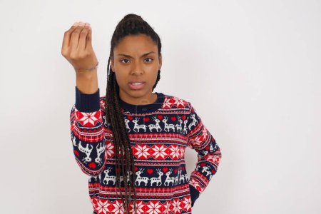 Photo for What the hell are you talking about. Shot of frustrated African American woman wearing Christmas sweater against white wall gesturing with raised hand doing Italian gesture, frowning, being displeased and confused with dumb question. - Royalty Free Image
