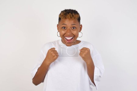 Téléchargez les photos : Jeune femme afro-américaine portant un t-shirt blanc se réjouissant de son succès et de la victoire serrant les poings avec joie.Lucky femme avec chapeau être heureux d'atteindre son but et ses objectifs.Émotions positives, sentiments. - en image libre de droit