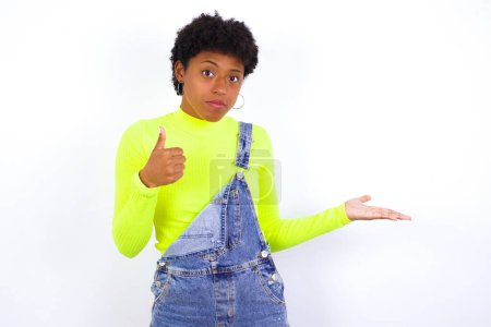 Photo for Happy cheerful young African American woman with short hair wearing denim overall against white wall showing thumb up and pointing with the other hand. I recommend this. - Royalty Free Image