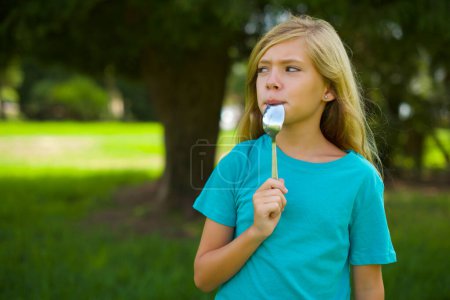 Porträt eines schönen kaukasischen kleinen Mädchens mit blauem T-Shirt, das draußen im Park steht und einen Löffel hält