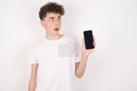 Téléchargez les photos : Beau jeune homme sur fond blanc tient un nouveau téléphone portable et semble mystérieux montre l'affichage vierge de cellulaire moderne - en image libre de droit