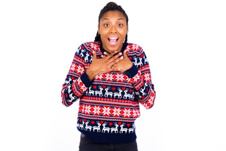 Foto de Feliz sonriente mujer afroamericana con suéter de Navidad contra la pared blanca tiene las manos en el pecho cerca del corazón. Emociones humanas, sentimientos reales y concepto de expresión facial. - Imagen libre de derechos