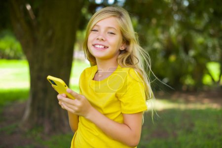 Téléchargez les photos : Petite fille caucasienne portant t-shirt jaune debout à l'extérieur Maquette espace de copie. Utilisation du téléphone mobile, dactylographier sms message - en image libre de droit