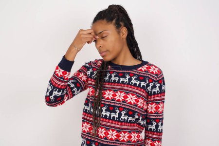 Foto de Mujer afroamericana muy molesta vistiendo suéter de Navidad contra pared blanca tocando la nariz entre los ojos cerrados, quiere llorar, tener una relación estresante o tener problemas con el trabajo - Imagen libre de derechos