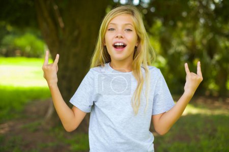 caucasian little kid girl wearing white t-shirt standing outdoor in the park makes rock n roll sign looks self confident and cheerful enjoys cool music at party. Body language concept.