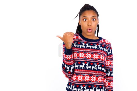 Téléchargez les photos : Choqué femme afro-américaine portant un pull de Noël contre les points de mur blanc avec le pouce loin, indique quelque chose. Regarde-moi ça. Concept de publicité. - en image libre de droit