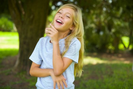 Photo for Portrait of caucasian little kid girl wearing white t-shirt standing outdoor in the park laughs happily keeps hand on chin expresses positive emotions smiles broadly has carefree expression - Royalty Free Image