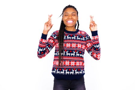 Téléchargez les photos : Joyeuse femme afro-américaine portant un pull de Noël contre un mur blanc serre les dents, lève les doigts croisés, fait un souhait souhaitable, attend de bonnes nouvelles, je dois gagner. - en image libre de droit