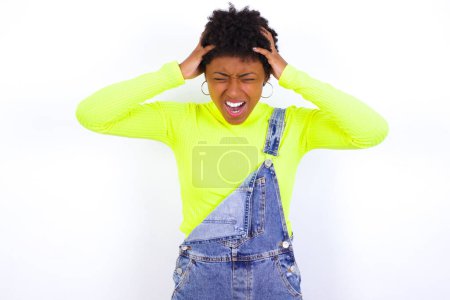 Photo for Shocked panic young African American woman with short hair wearing denim overall against white wall holding hands on head and screaming in despair and frustration. - Royalty Free Image