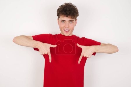 Foto de Joven hombre guapo vistiendo camiseta roja con expresión positiva, apunta hacia abajo con ambos dedos índice, tiene amplia sonrisa interesada. Mira ahí, por favor.. - Imagen libre de derechos