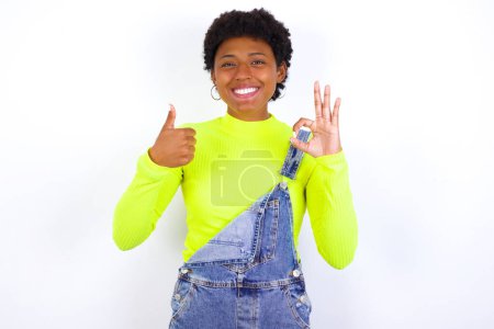 Téléchargez les photos : Jeune afro-américaine aux cheveux courts portant globalement du denim contre un mur blanc souriant et regardant heureux, insouciant et positif, geste de victoire ou de paix d'une main - en image libre de droit